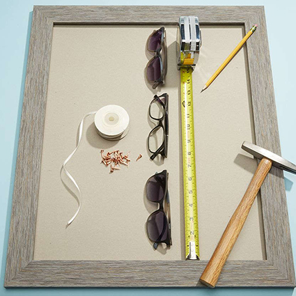 Gray frame filled with glasses, a measuring tape, a pencil, a hammer, ribbon, and pencil shavings.