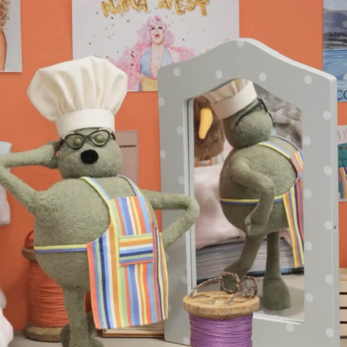 Green puppet wearing a chef's hat, glasses, and a colorful striped apron poses confidently in front of a mirror.