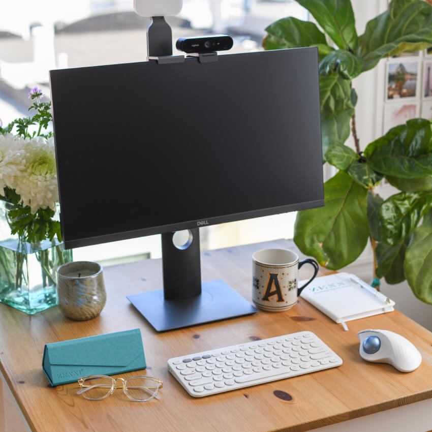 Dell monitor, webcam, wireless white keyboard, mouse, mug with letter "A", glasses, and teal notebook on desk.