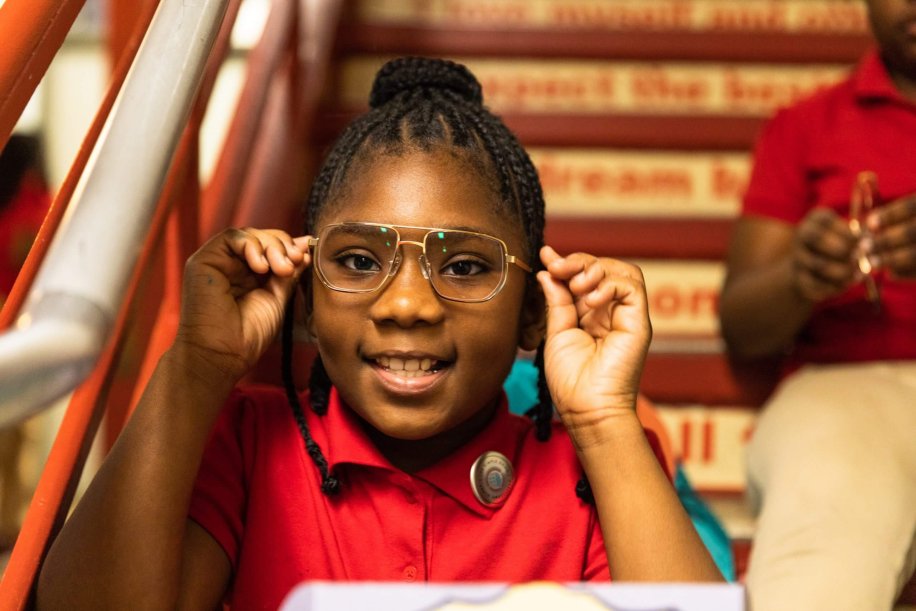 Child wearing large glasses, smiling. Background includes blurred text on steps: "Dream the impossible."