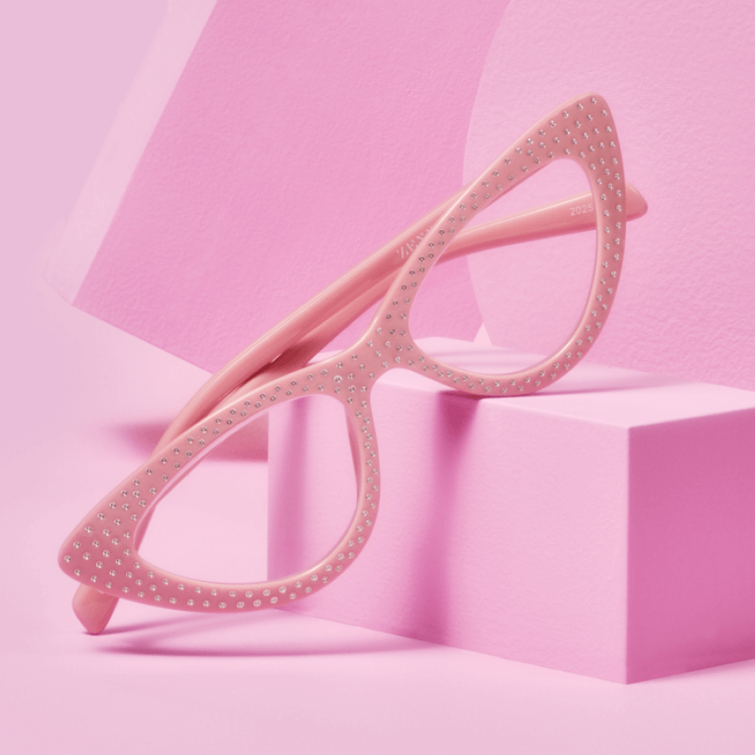 Pink cat-eye glasses with stud details on the frame, set against a pink geometric background.