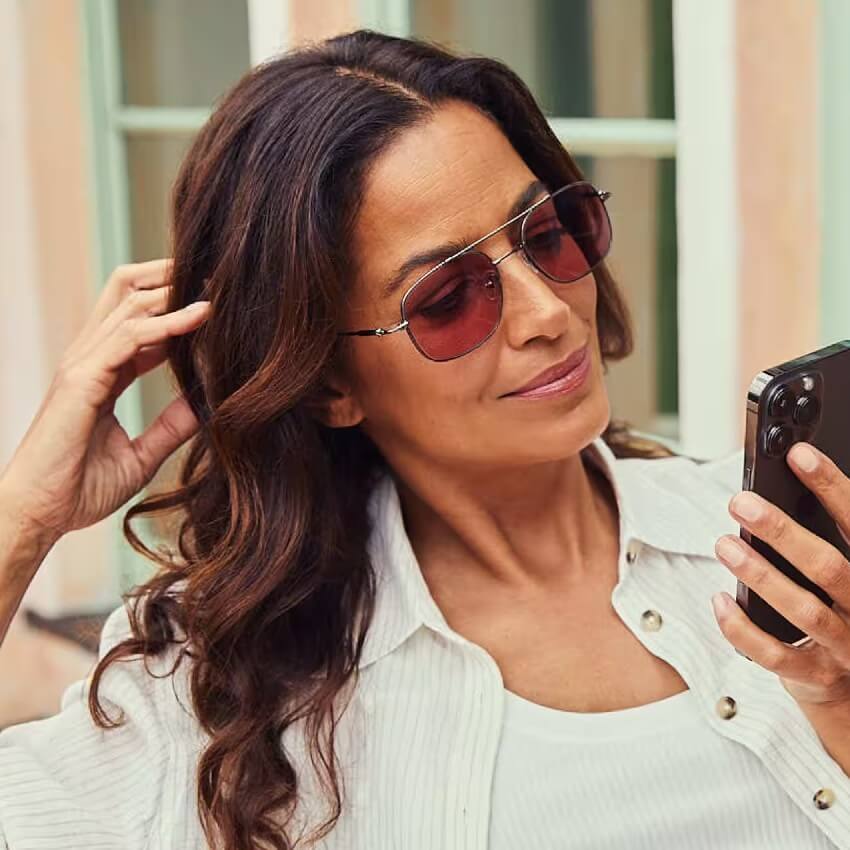 Woman wearing aviator sunglasses with pink-tinted lenses, looking at a phone.