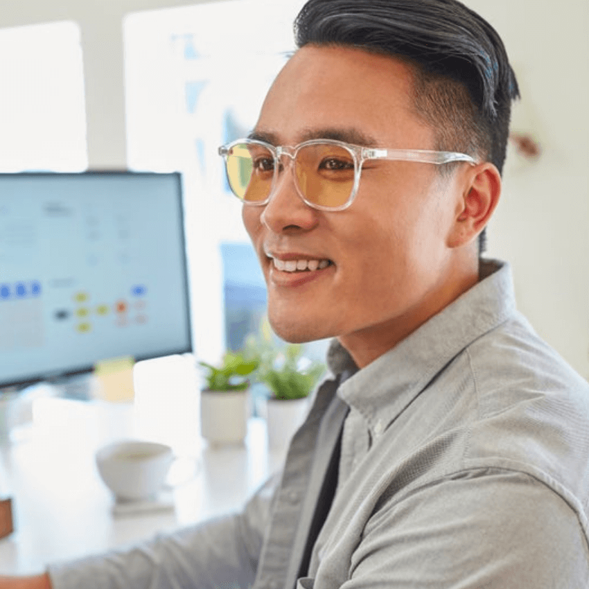 Man wearing clear frame, orange-tinted glasses, smiling and working at a desk with a computer monitor.
