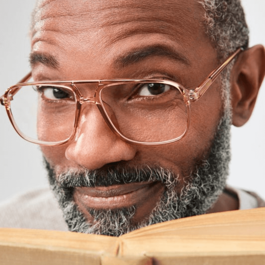 Man wearing large, clear-framed glasses, looking over an open book.