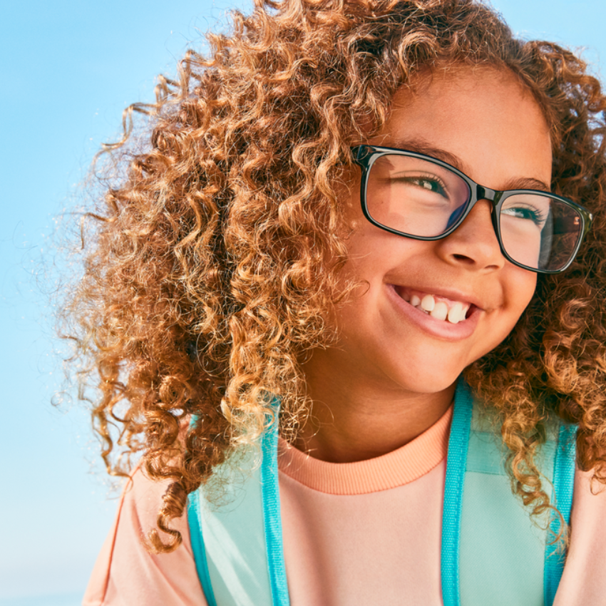 Smiling child with curly hair wearing black-rimmed glasses and a teal backpack.