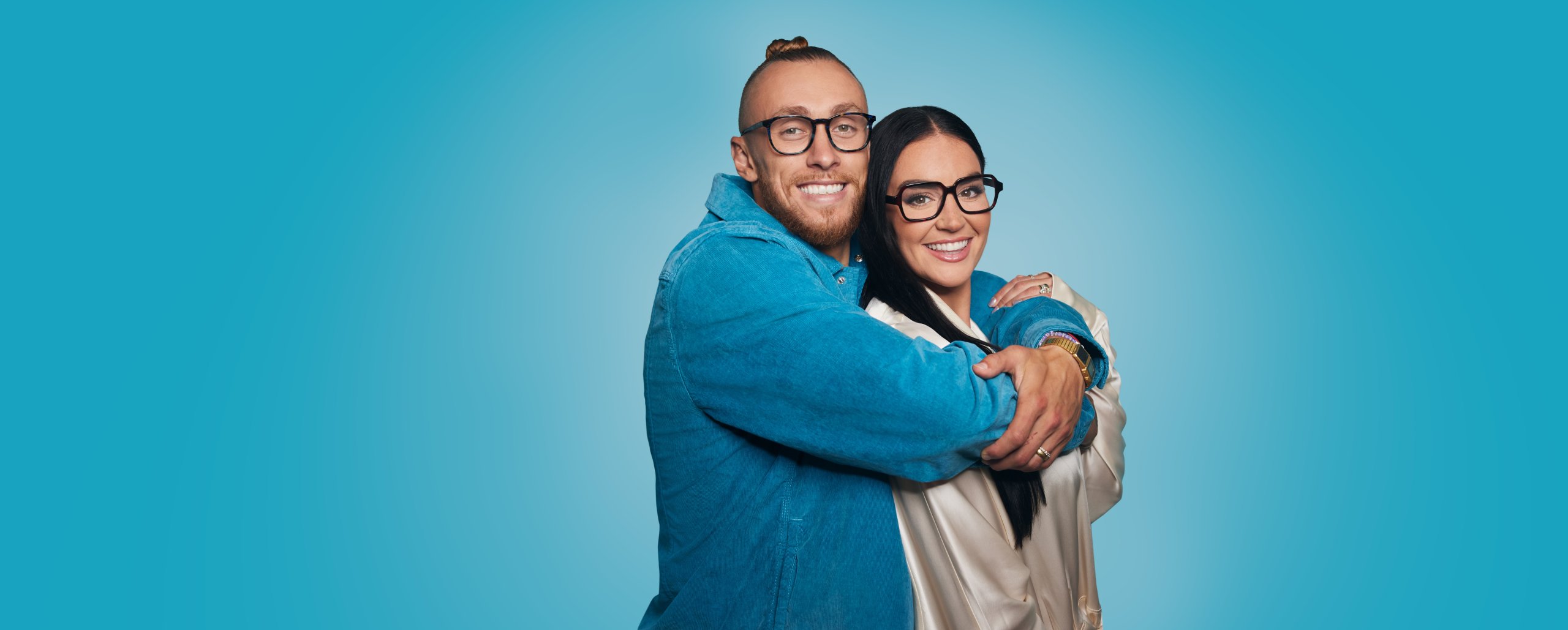 Two people happily embracing, both wearing eyeglasses and smiling against a blue background.