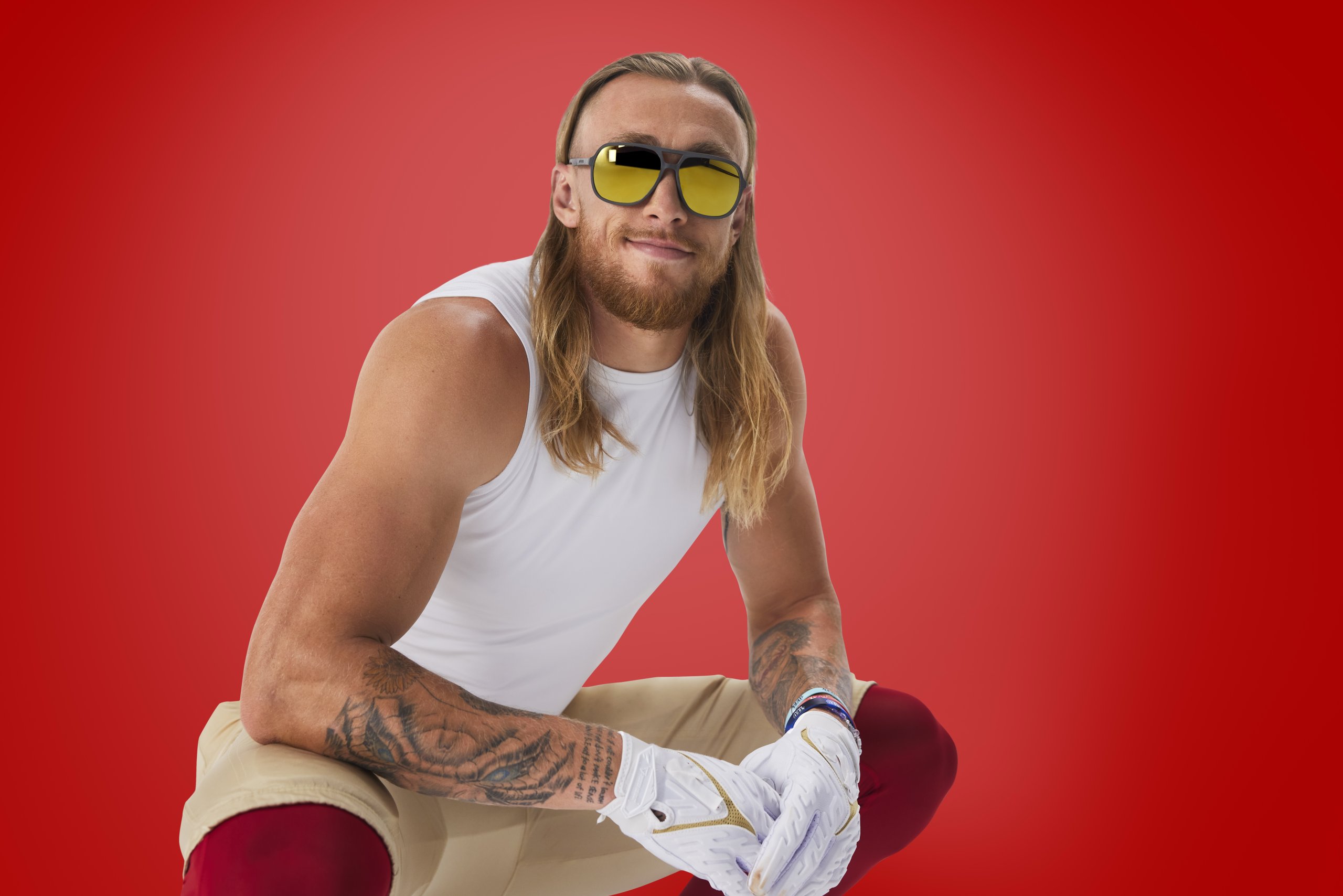 Man wearing sunglasses, white sleeveless shirt, white gloves, and beige-and-red pants on a red background.