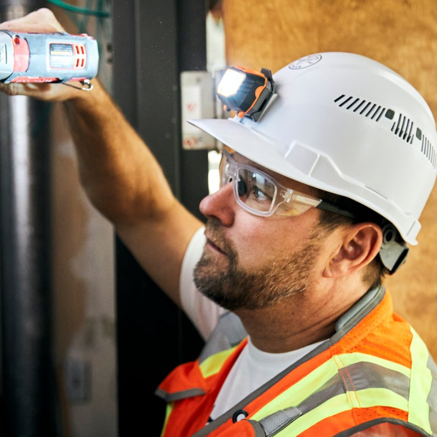 Worker in safety gear using a tool, wearing a white helmet with a headlamp, clear glasses, and an orange vest.