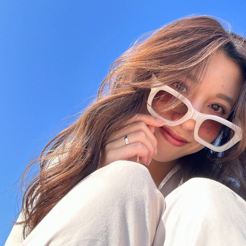 Woman wearing oversized white sunglasses and a light-colored outfit, smiling with a clear blue sky background.