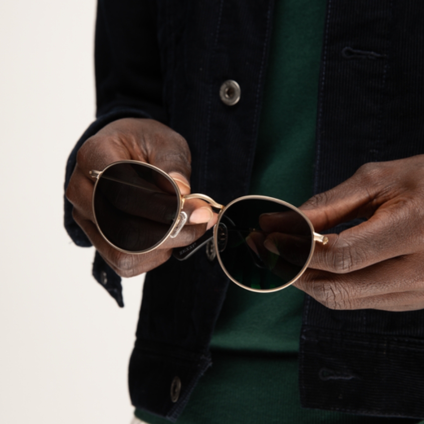 Hands holding gold-framed round sunglasses with dark lenses.