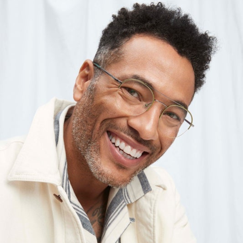 Man wearing thin, round-frame glasses with a wide smile and a light-colored collared jacket.