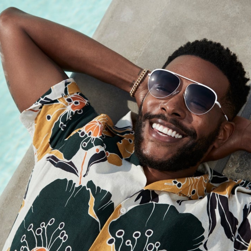 Man with a floral Hawaiian shirt and aviator sunglasses smiling, reclining by a pool.