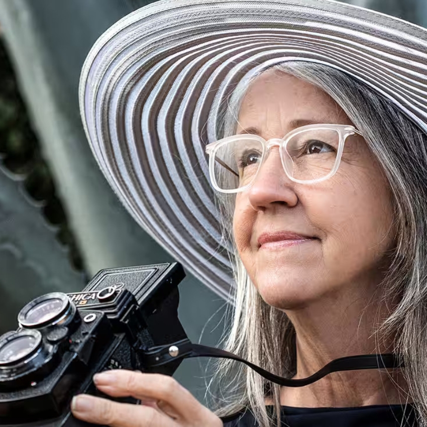A person holding a Yashica-D twin-lens reflex camera, wearing a white-striped wide-brimmed hat and glasses.