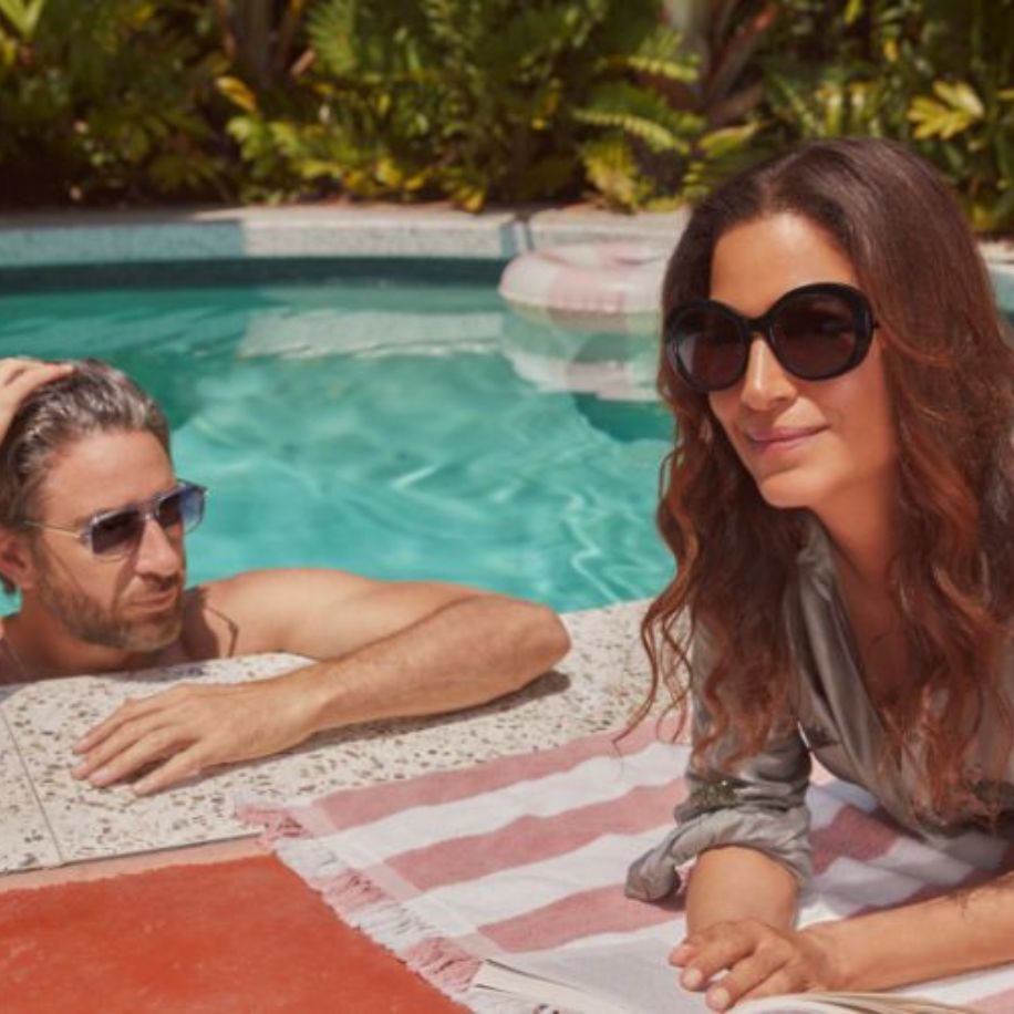 Man in sunglasses in pool and woman in large black sunglasses lying on a striped towel by the pool.