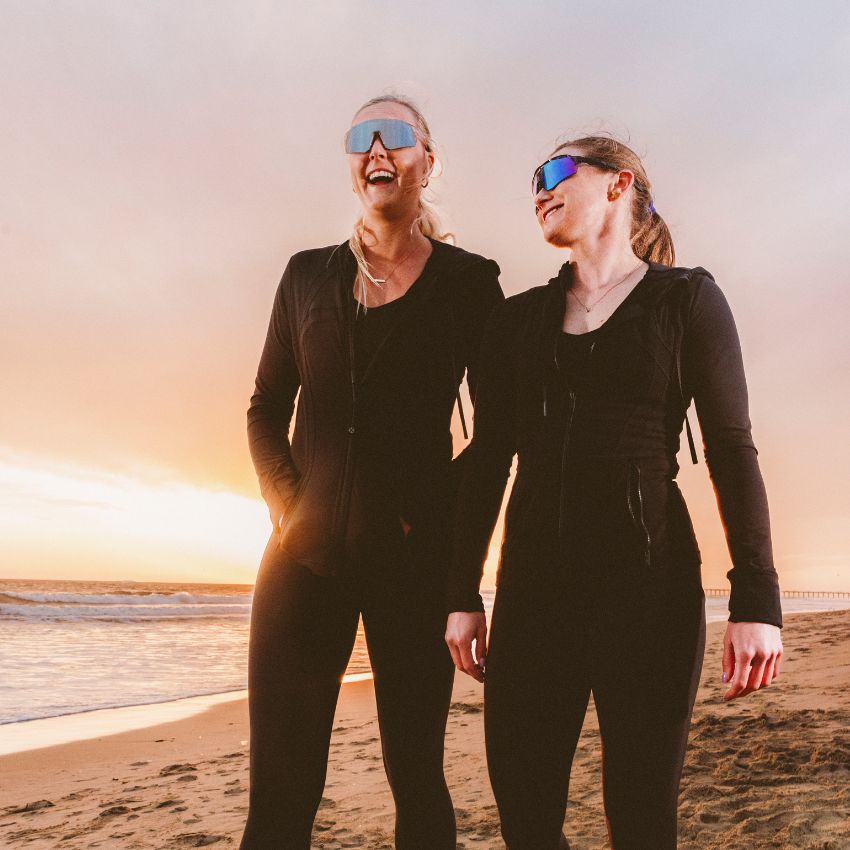 Two women wearing black activewear and reflective sunglasses standing on a beach at sunset.